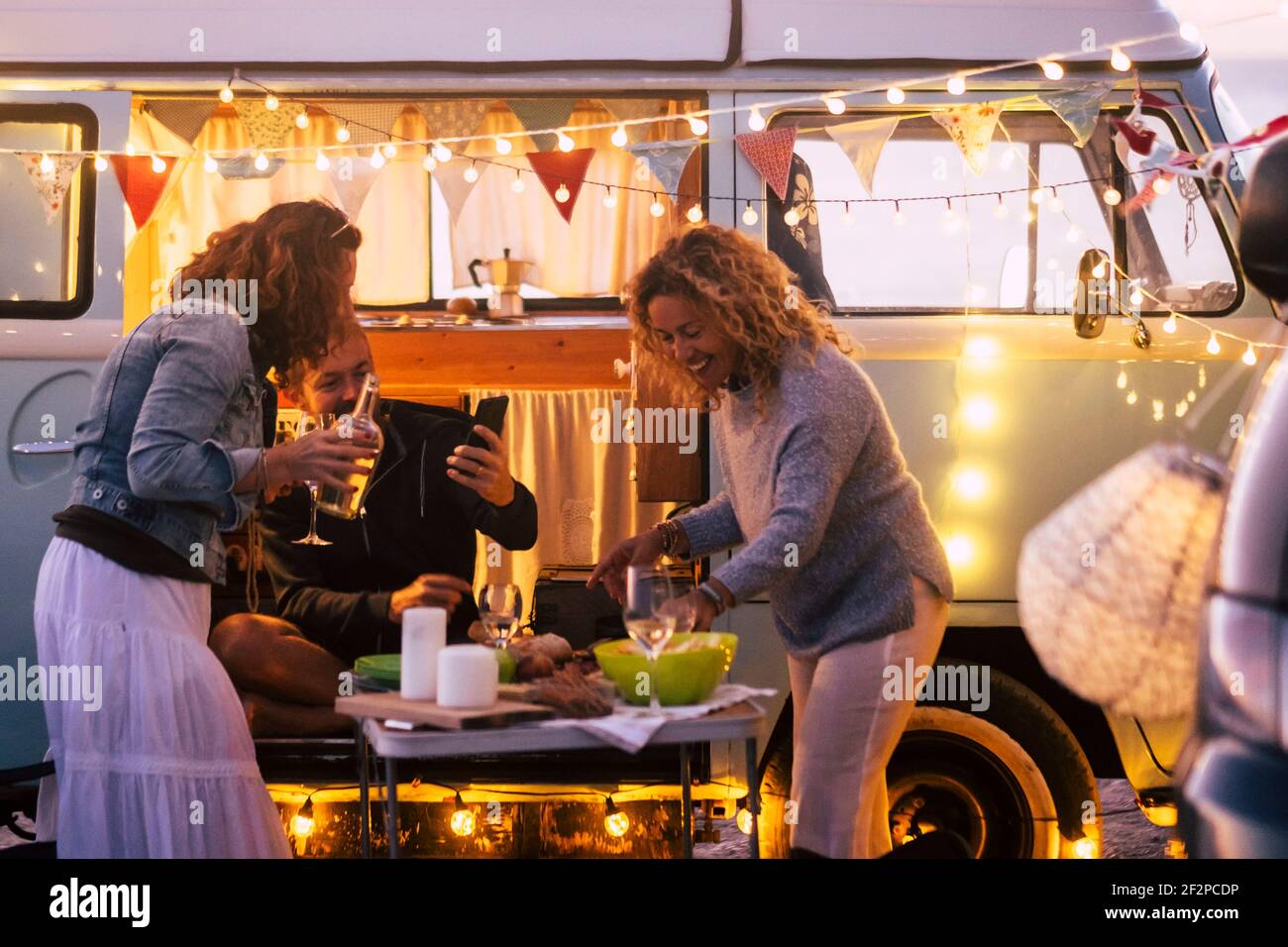 Gruppe von drei erwachsenen Menschen Freunde feiern und essen zu Abend Gemeinsam im Outdoor-Urlaub mit Tafelkost und Vintage Van im Hintergrund - alternativer Lebensstil und Glück Stockfoto