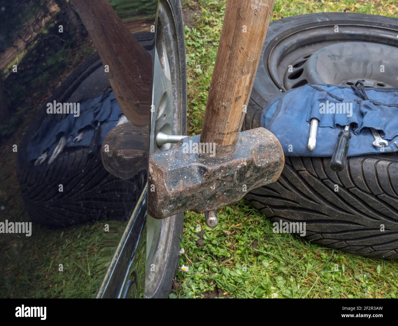 Ein Vorschlaghammer trifft auf eine Radhalterung / einen Schraubenschlüssel auf einem Fahrzeug, um eine Radmutter zu lösen, mit einem Reserverad und einem Werkzeugsatz in der Nähe. Stockfoto