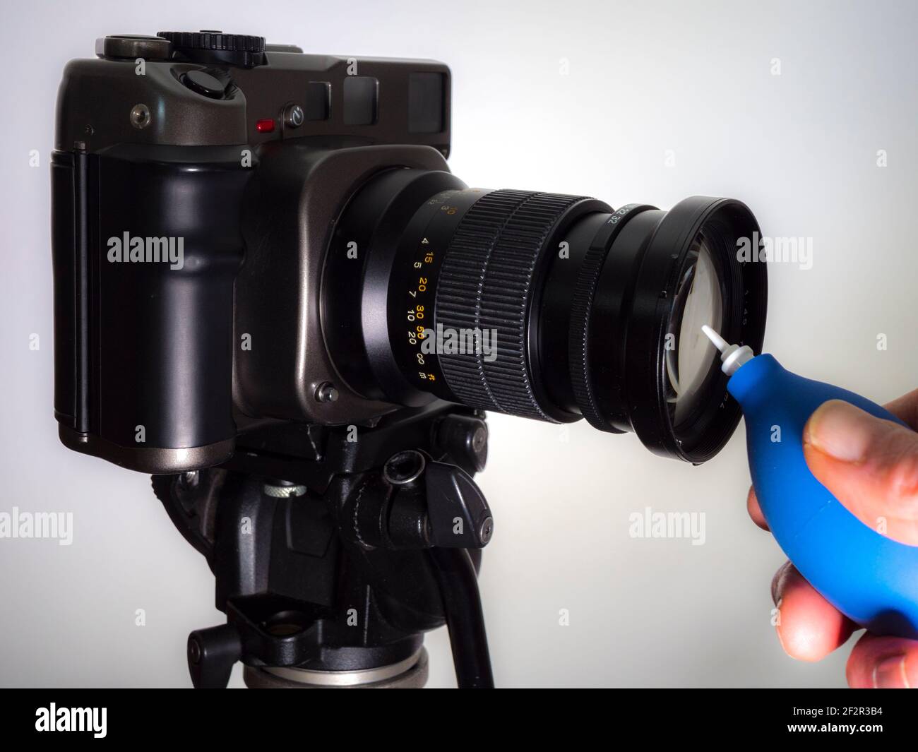 Nahaufnahme isolierte POV-Aufnahme einer Männerhand mit einem blauen Gummibläser, um Staub aus dem Glas eines Mittelformat-Filmkameraobjektivs auf einem Stativ zu entfernen. Stockfoto