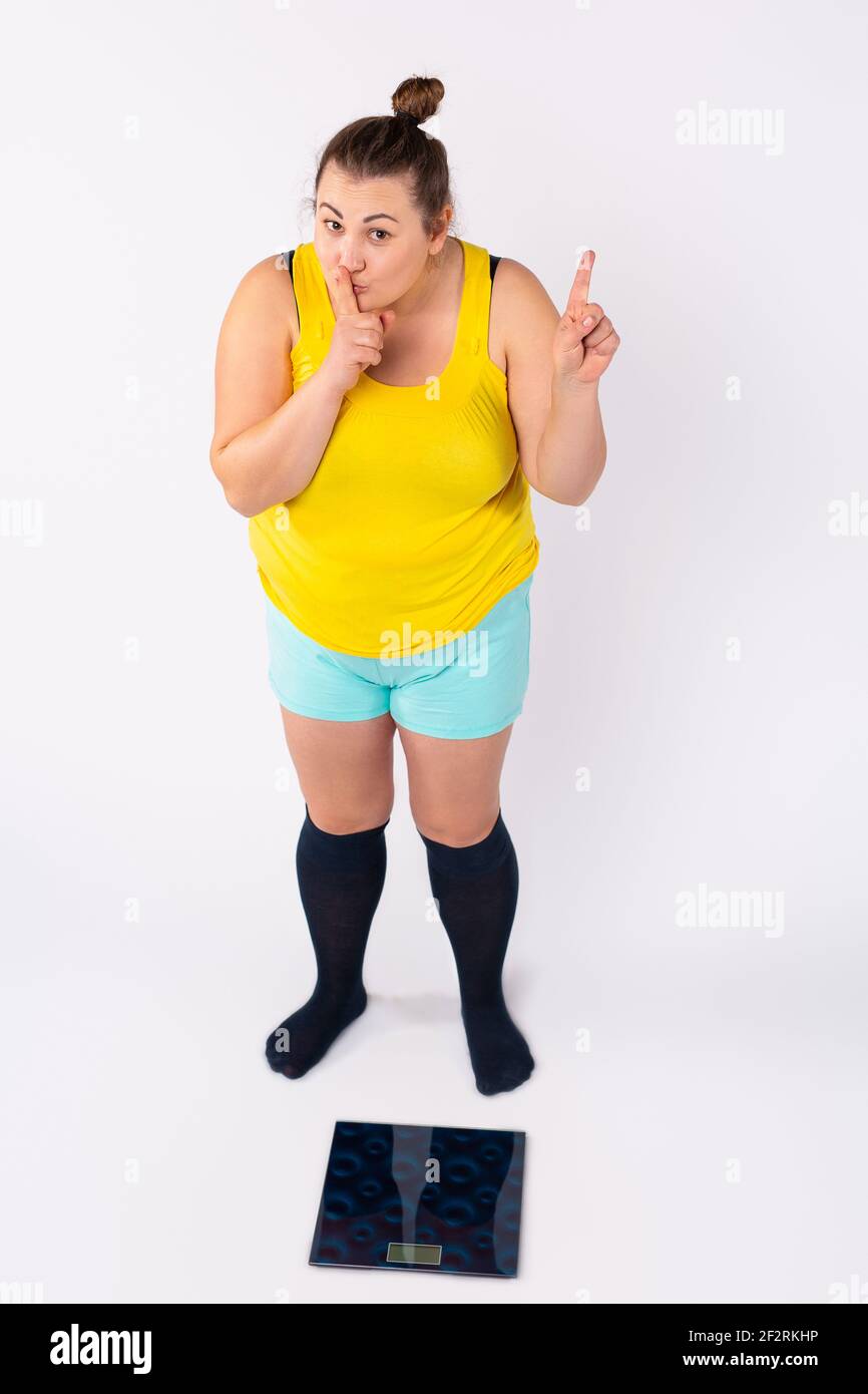 Eine kaukasische prall gekleidete Frau steht vor einer elektronischen Waage und zeigt ein Schild, das ihren Zeigefinger leise vor ihr Gesicht hält. Stockfoto