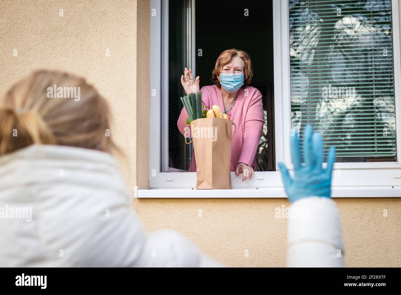 Soziale Distanzierung durch Coronavirus covid-19 Pandemiesperre. Ältere Frau mit Gesichtsmaske winkte aus dem Fenster zu ihrer erwachsenen Tochter, sie lieferte Essen Stockfoto