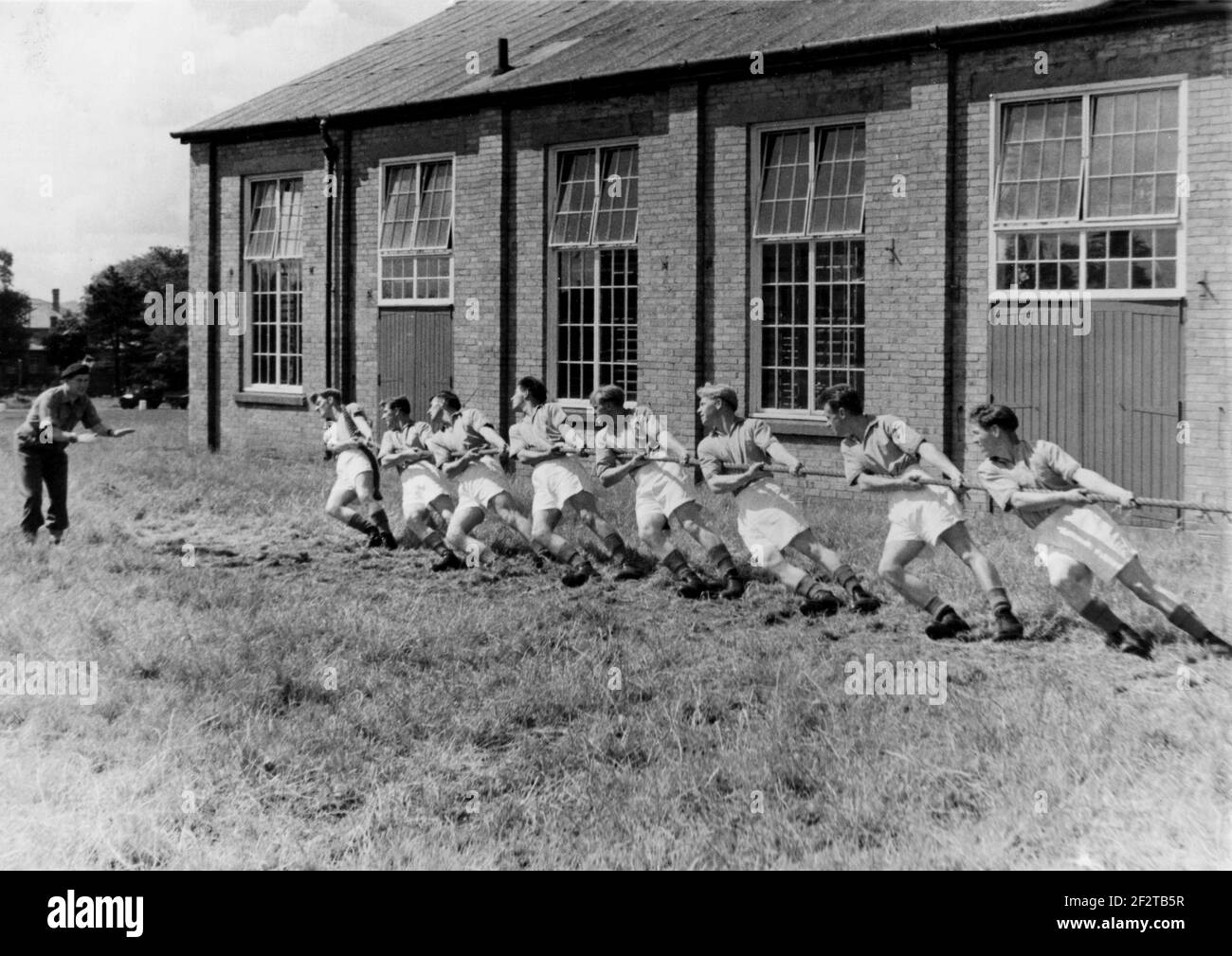 1950s, historischer, nationaler Dienst nach dem Krieg, ein Armeeoffizier, der acht junge Soldaten in ihren Turnschuhen und Armeestiefeln Anweisungen gibt, die bei einer Tauziehen-Übung auf Gras vor ihren Kasernen, England, Großbritannien, an einem Seil ziehen. Stockfoto