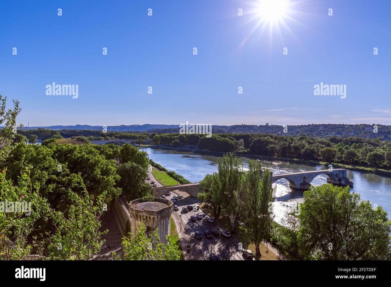 Die Pont Saint-Benezet erstreckt sich über die Rhône in Avignon, Frankreich, gesehen über baumgesäumten Ufern und Stadtmauern unter einem hellen Sonnenstrahl. Stockfoto
