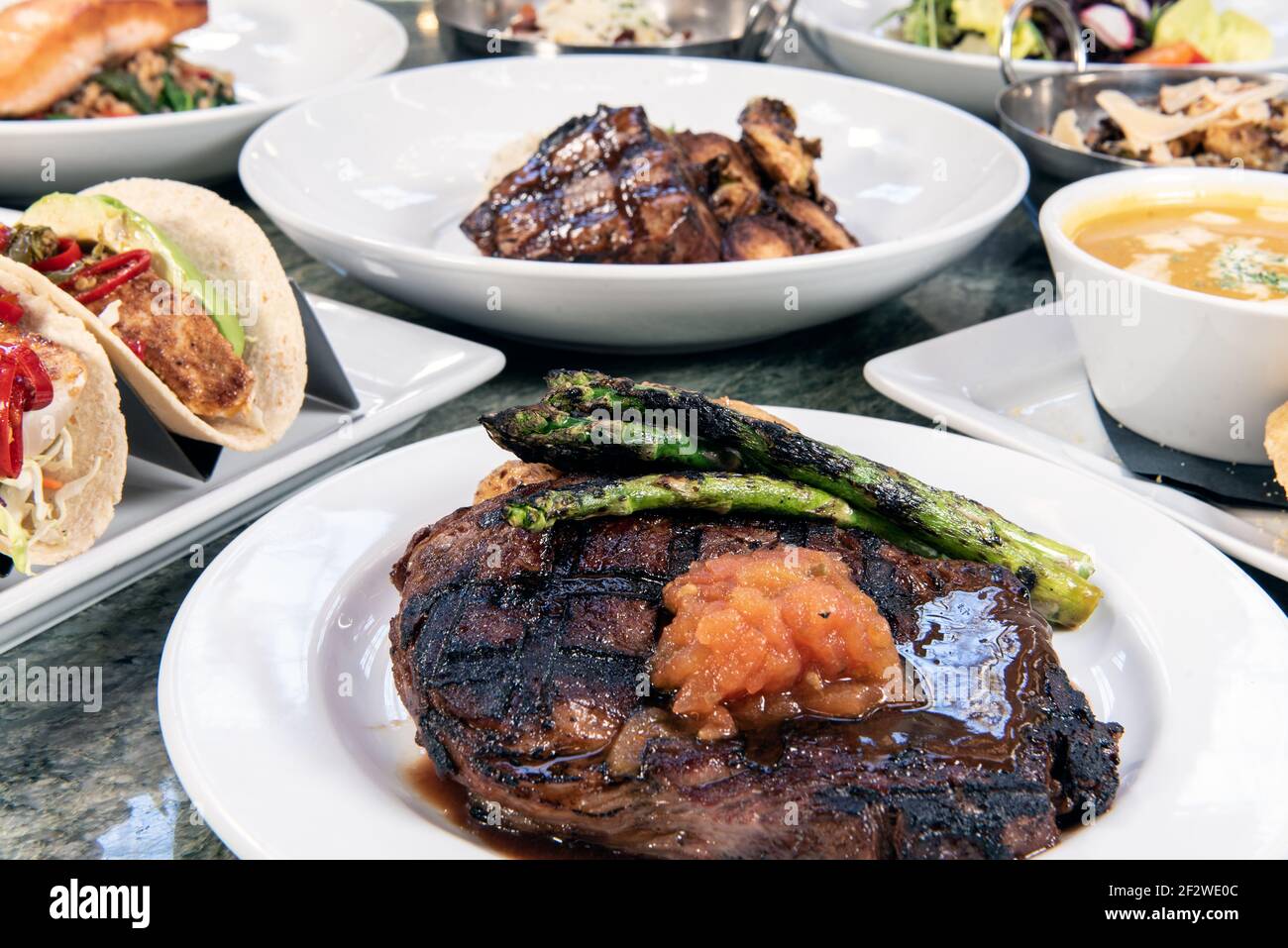 Voller Tisch mit einer Vielfalt an Restaurantgerichten zur Auswahl mit Rib Eye Steak in der Mitte. Stockfoto