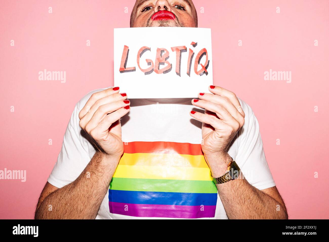 Rebellischer bärtiger homosexueller Mann mit roten Lippen und Maniküre machen Grimace beim Anzeigen und Bedecken Gesicht mit Papier mit LGBTIQ Text gegen rosa b Stockfoto