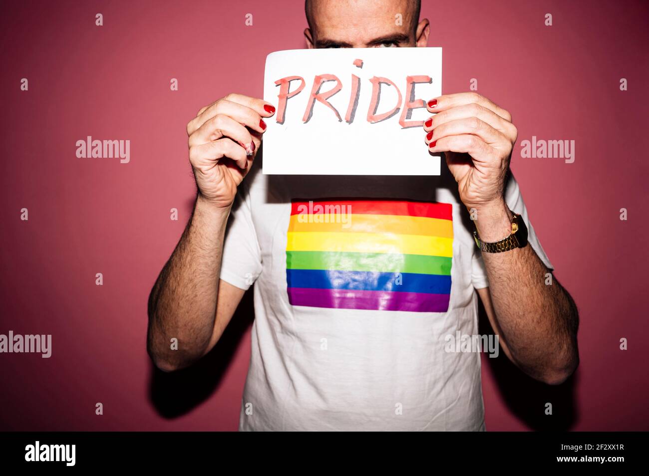 Rebellischer bärtiger homosexueller Mann mit roten Lippen und Maniküre machen Grimace beim Zeigen und Bedecken Gesicht mit Papier mit Pride Text gegen rosa BA Stockfoto