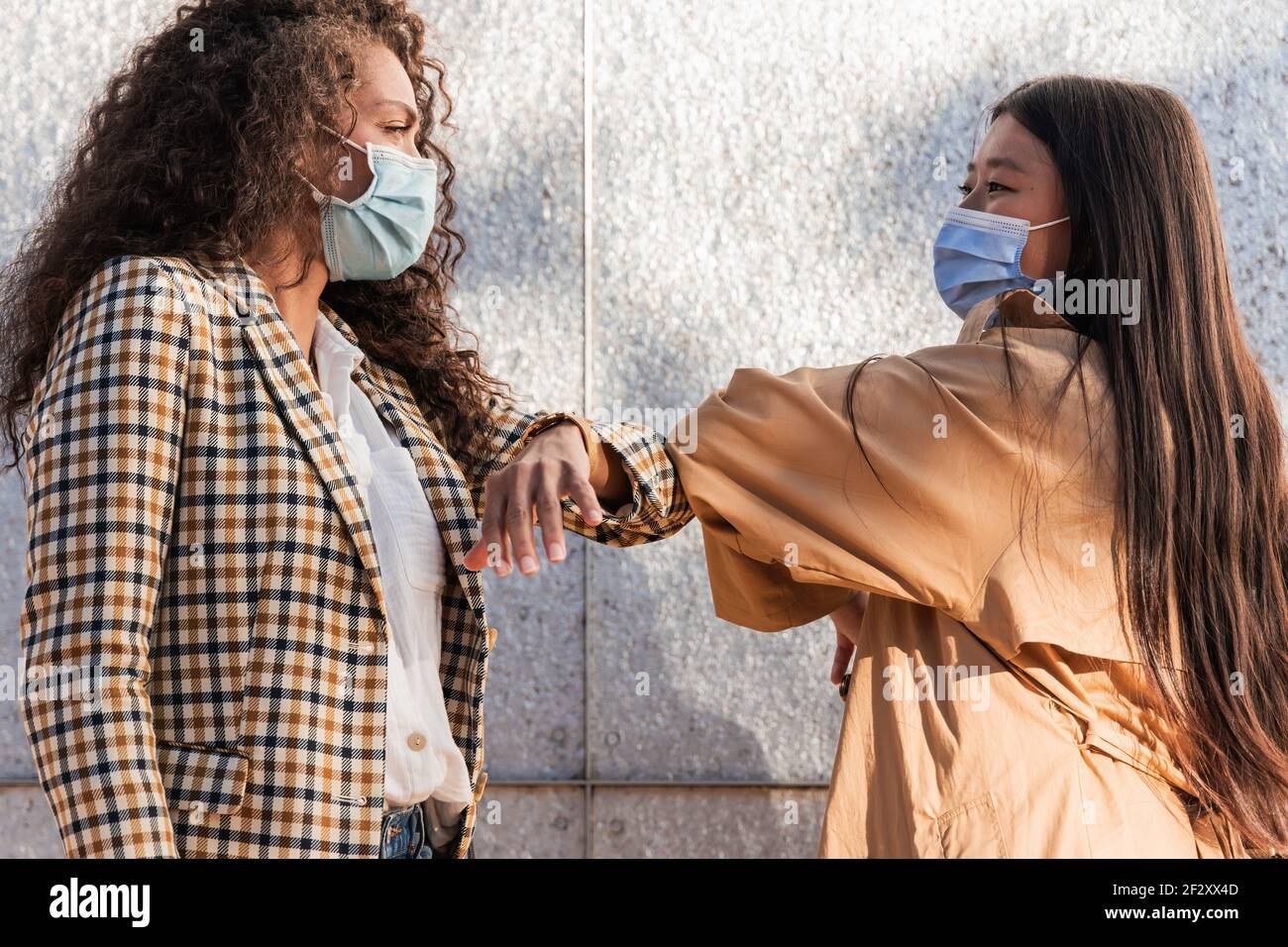 Seitenansicht der ethnischen weiblichen Freunde in legerer Kleidung mit Medizinische Masken, die einander anblicken, während sie mit Ellbogen grüßen Stockfoto