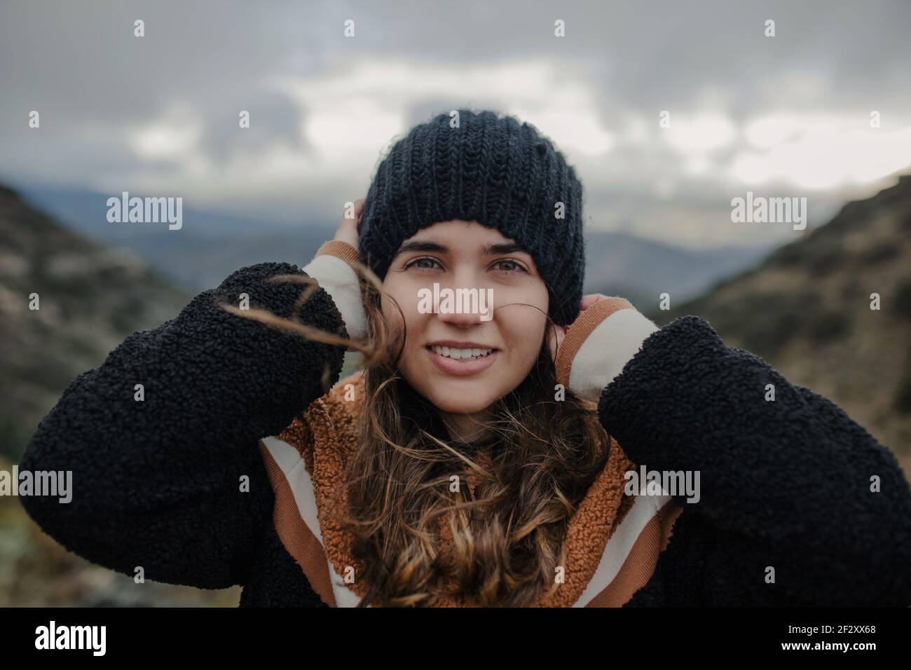 Lächelnde junge Frau, die warmen Hut aufsetzt und anschaut Kamera beim Stehen auf rauem Hochland an wolkig düsteren Tag Stockfoto