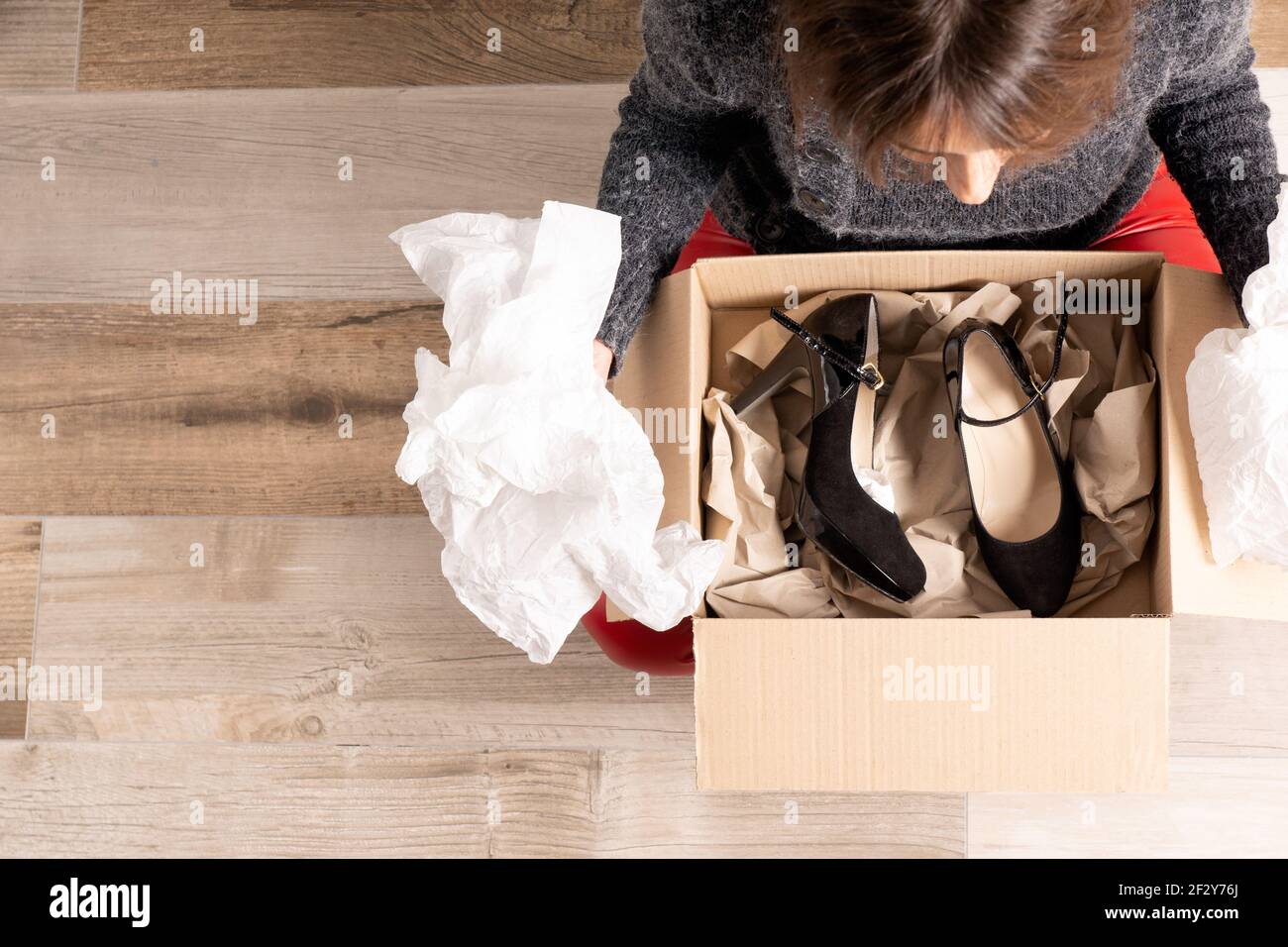 Schuss von oben von jungen Mädchen in roten Hosen sitzen Auf dem Holzboden ein Paket zu öffnen, das sie gerade gekauft hat Online mit einigen schwarzen High Heels Stockfoto