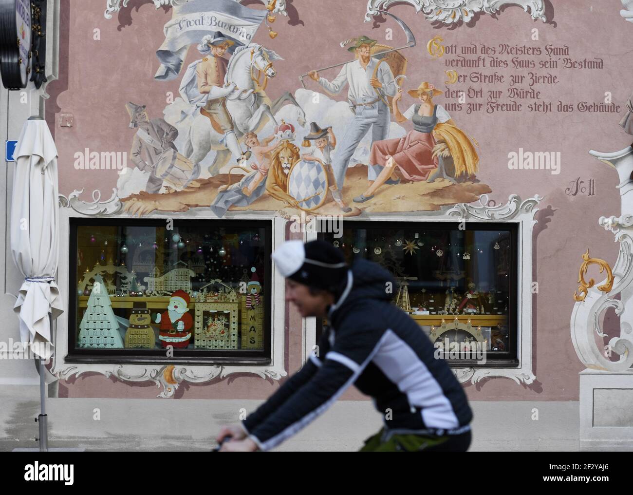 Oberammergau, Deutschland. Februar 2021, 17th. Ein Mann mit FFP2 Masken fährt an einer Holzschnitzerei vorbei, die wegen der Korona geschlossen ist, Adventskalender sind noch im Schaufenster zu sehen. Das Geschäft mit Weihnachtskrippen, aber vor allem mit Kruzifixen, läuft das ganze Jahr über - aber die wichtige Vorweihnachtszeit und vor allem die Passion wurden 2020 wegen der Pandemie gestrichen. Quelle: Angelika Warmuth/dpa/Alamy Live News Stockfoto