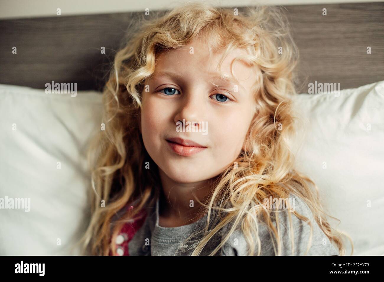 Ziemlich schöne kaukasische blonde Mädchen mit langen unordentlichen Haaren und blauen Augen sitzen auf einem Bett zu Hause Blick auf die Kamera. Nachdenkliche Kind Kind Traum denken Stockfoto