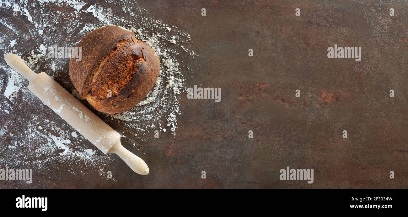 Hausgemachtes Sauerteigbrot mit, Nudelholz und verschüttetem Mehl auf einem dunklen Küchentisch mit Kopierplatz. Stockfoto