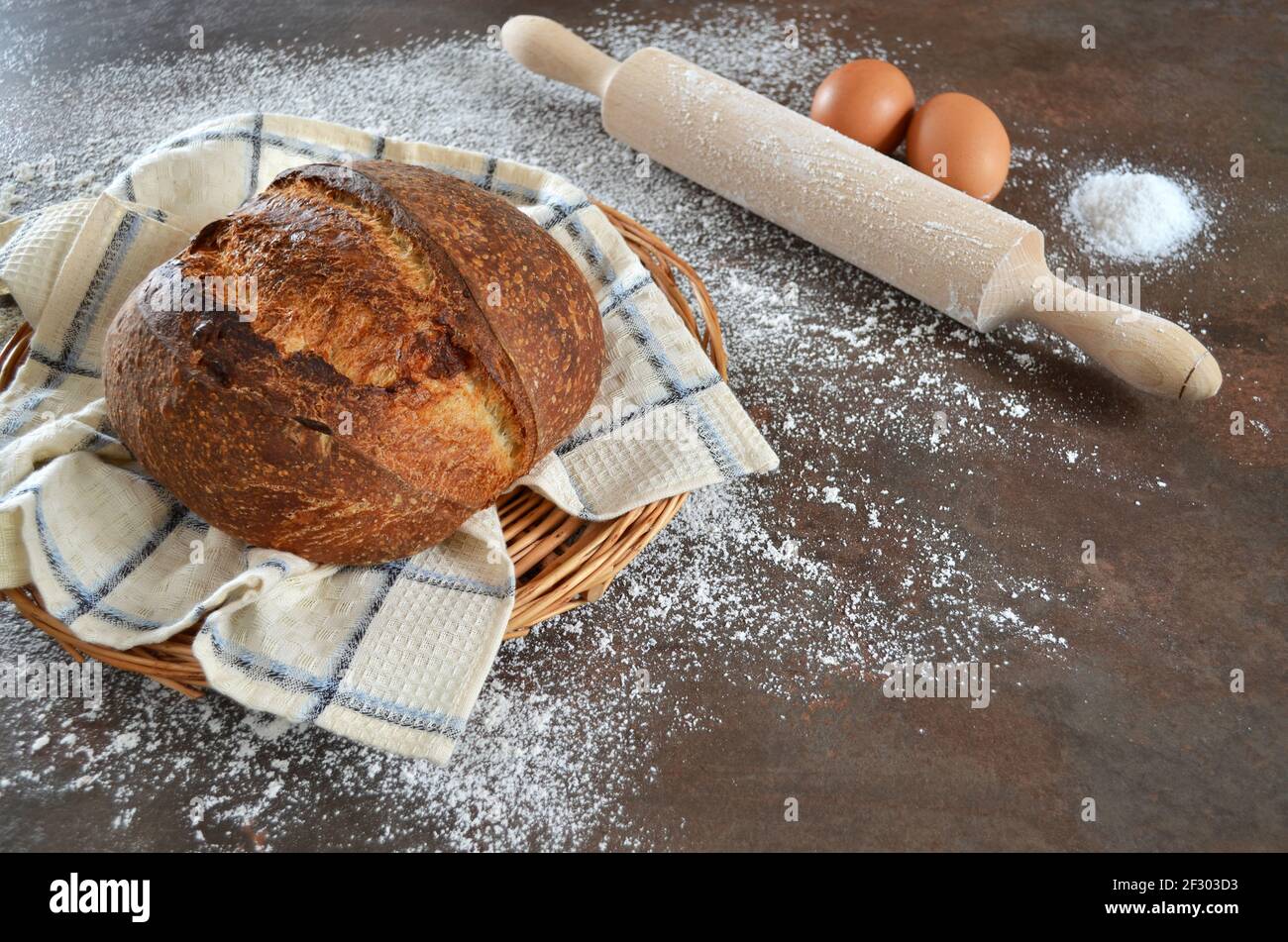 Frisch gebackenes selbstgebackenes Sauerteig-Brot mit einer knusprigen Kruste auf einem Küchentuch auf dem Küchentisch mit Mehl bestreut. Ein Nudelholz, zwei Eier und ein Stockfoto