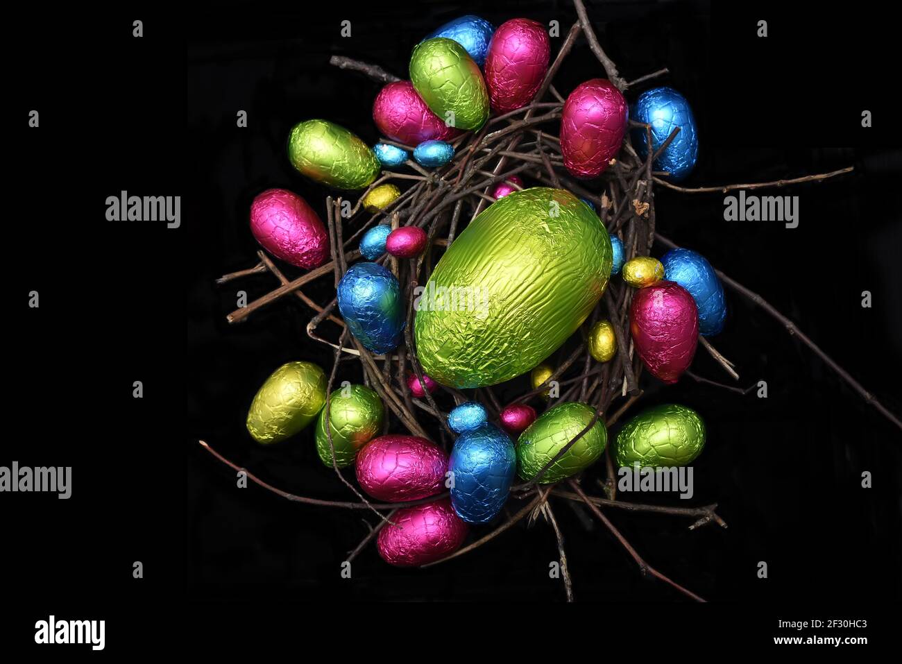 In Folie verpackte mehrfarbige ostereier in rosa, grün, blau und gelb in einem natürlichen Nest aus Holz, Stäbchen und Zweigen, vor schwarzem Hintergrund. Stockfoto