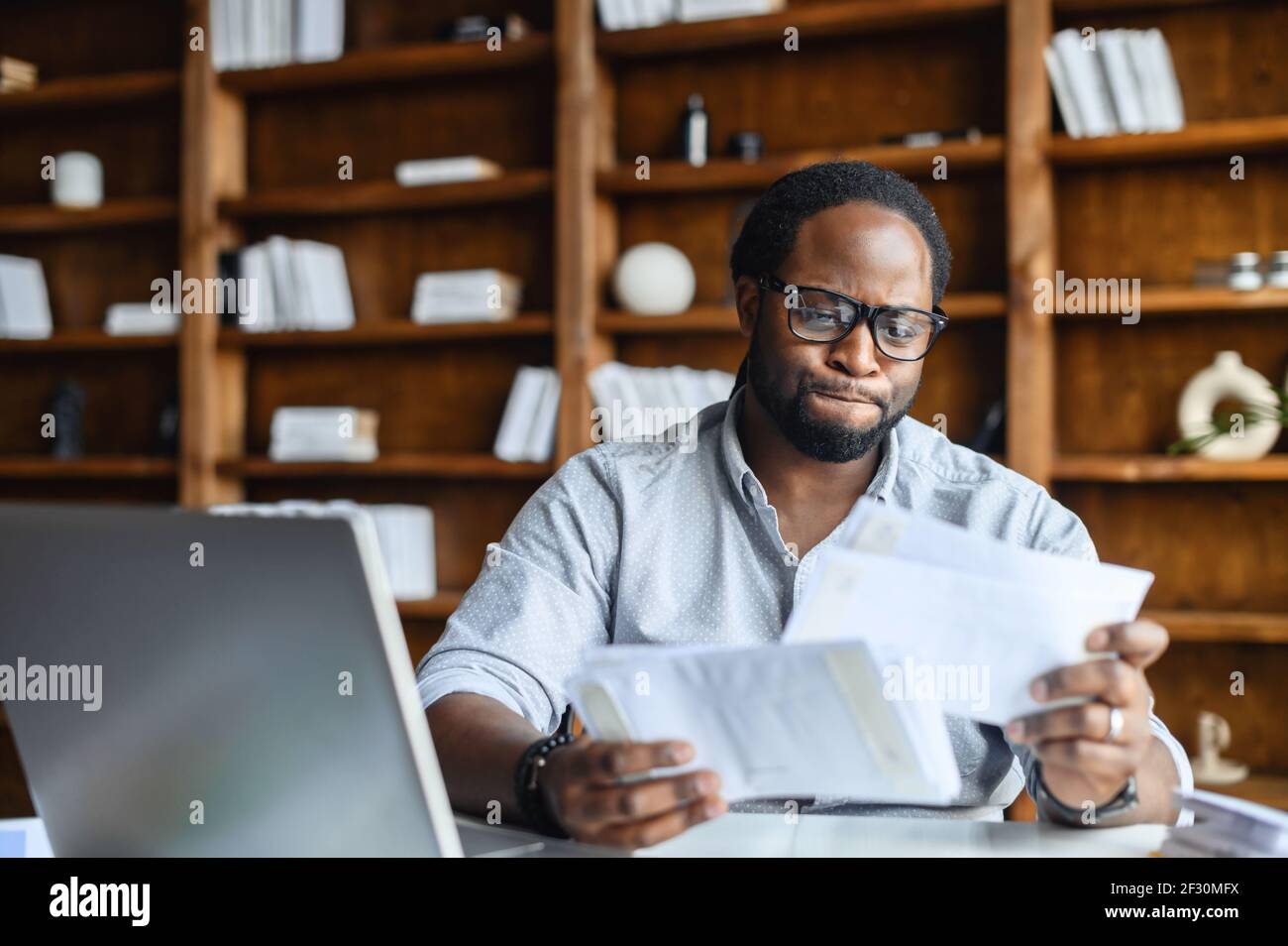 Verwirrt intelligente junge afroamerikanische Student oder Unternehmer in einer Brille überprüfen Zahlen aus Papieren, sitzen am Schreibtisch in einer Bibliothek und halten Dokumente, Korrekturlesen Vereinbarung mit dem Kunden Stockfoto