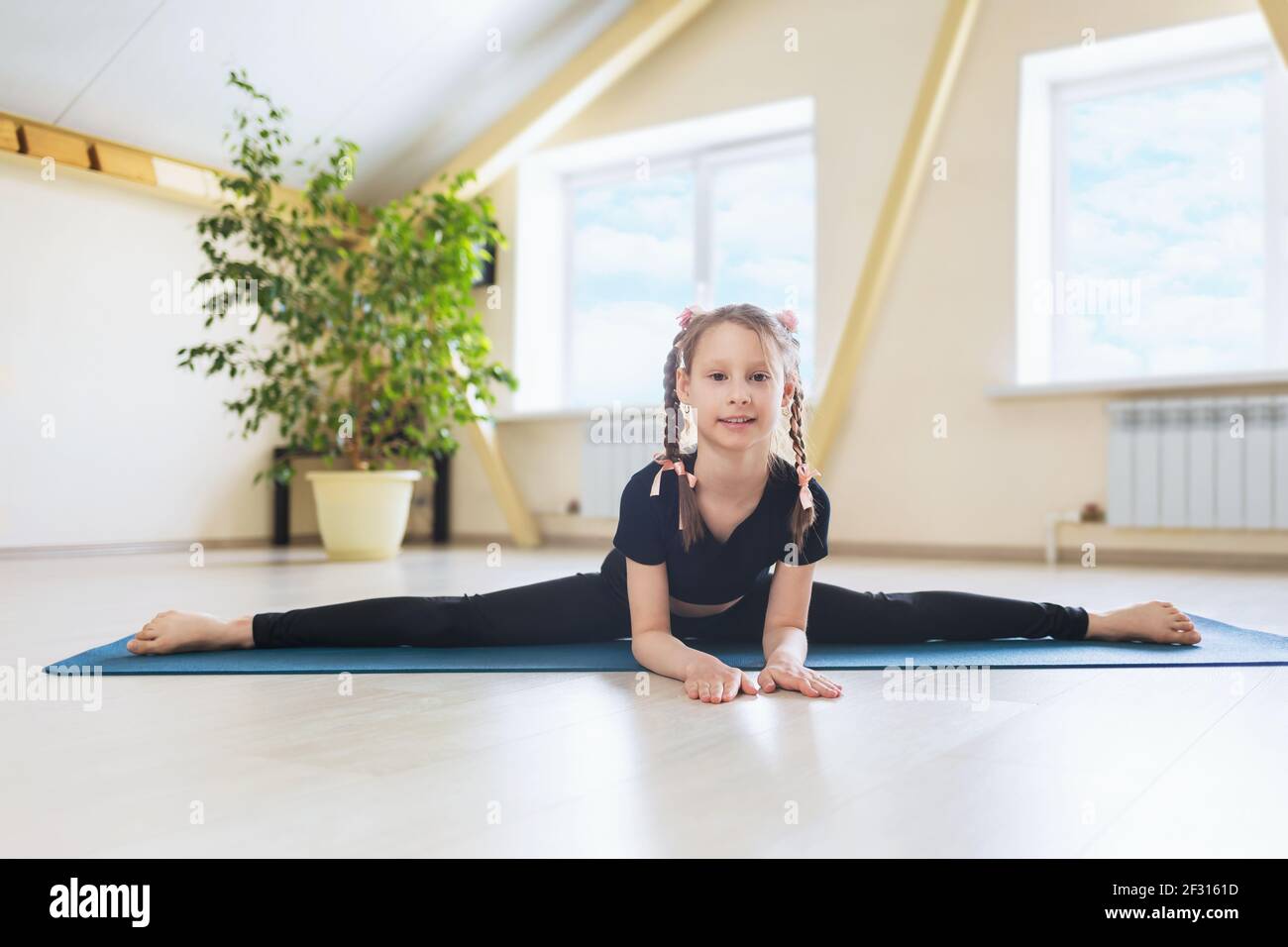 Ein glückliches kleines Mädchen in schwarzer Sportkleidung, das Yoga praktiziert, führt einen Querspalt auf einer Gymnastikmatte, der Samokanasana Übung, durch. Stockfoto