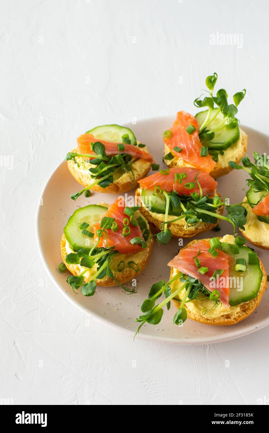 Leckere herzhafte Vorspeise, Lachsbraten auf einem weißen Teller, rundes gebratenes Brötchen mit rotem Fisch, Gurke, Sauce Hollandaise und Erbsensprossen Stockfoto