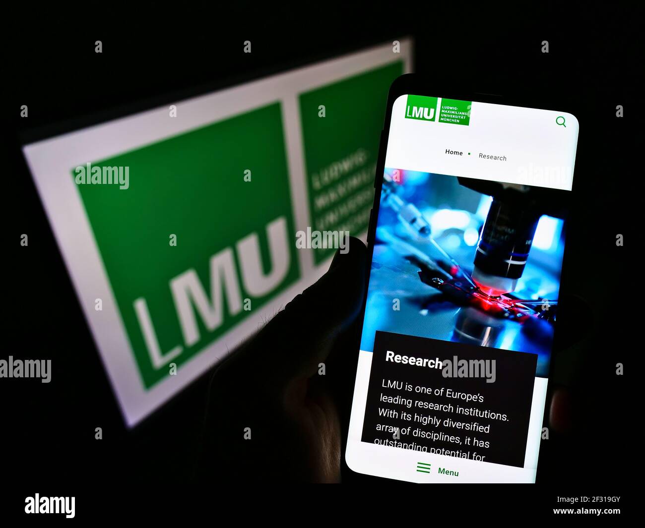 Person, die Handy mit Webseite der LMU München auf dem Bildschirm vor dem Logo hält. Konzentrieren Sie sich auf die Telefonanzeige. Nicht modifiziertes Foto. Stockfoto