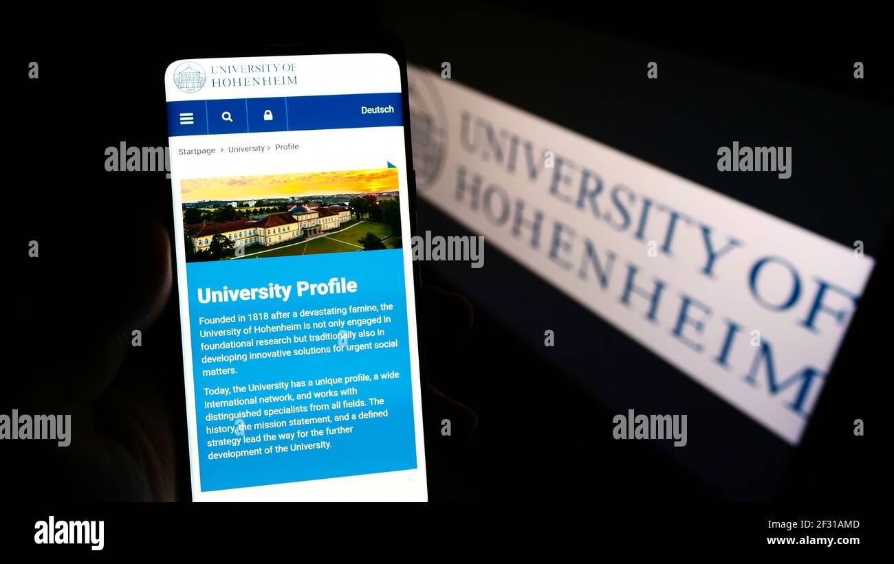 Person, die Mobiltelefon mit Website der Universität Hohenheim auf dem Bildschirm vor dem Logo hält. Fokus auf der Mitte des Telefondisplays. Stockfoto