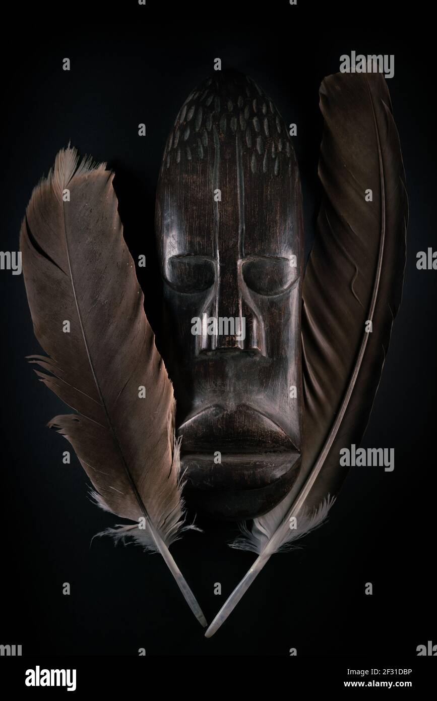 Holzskulptur des menschlichen Gesichts im afrikanischen Stil mit großen Federn um ihn herum. Holzschnitzerei aus dunklem Holz. Afrikanische Stammesmaske isoliert auf schwarzem Hintergrund. Kunst Stockfoto
