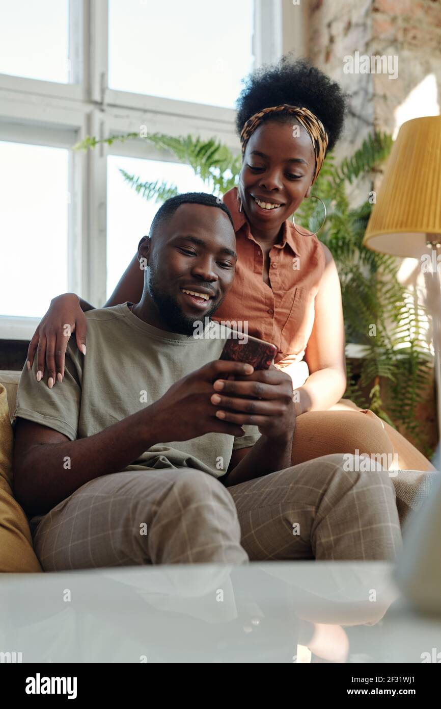 Fröhliche junge afrikanische Paar in Casualwear Blick auf Smartphone-Bildschirm Mit einem Lächeln, während Sie vor der Kamera gegen große sitzen Angezeigt Stockfoto
