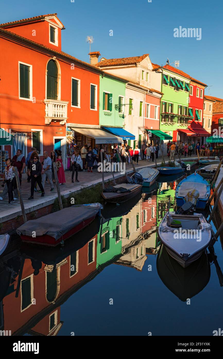 Die venezianische Insel Burano, Geschäfte mit Menschen, die entlang eines Kais neben einem Kanal mit Reflexionen der Szene spazieren. Stockfoto