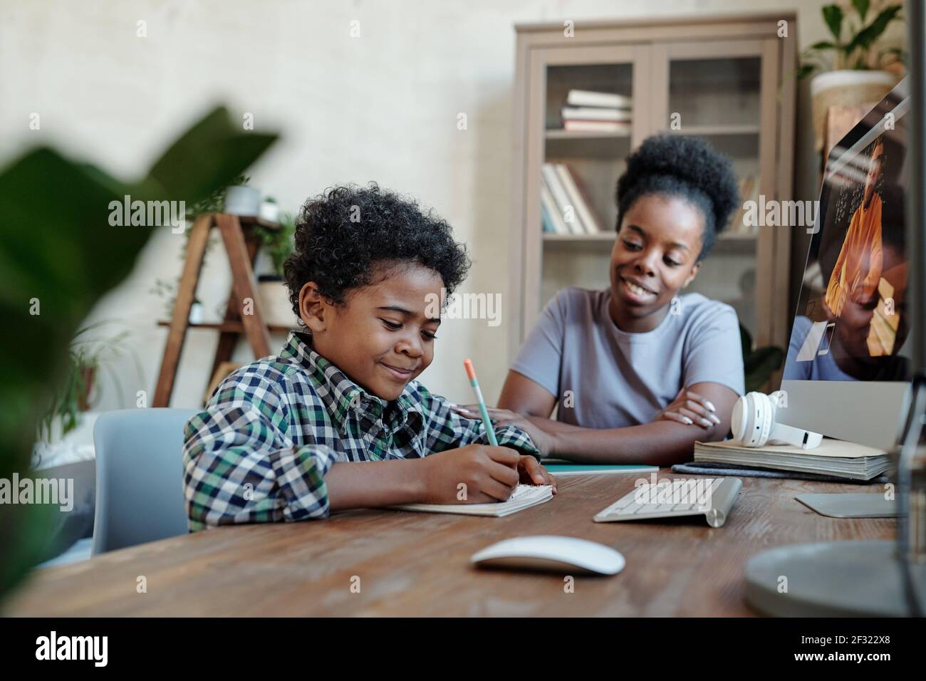 Fleißiger Schuljunge macht Notizen vor dem Monitor mit seinem Lehrer auf dem Bildschirm während des Online-Unterrichts, während seine Mutter sitzt Neben ihm Stockfoto