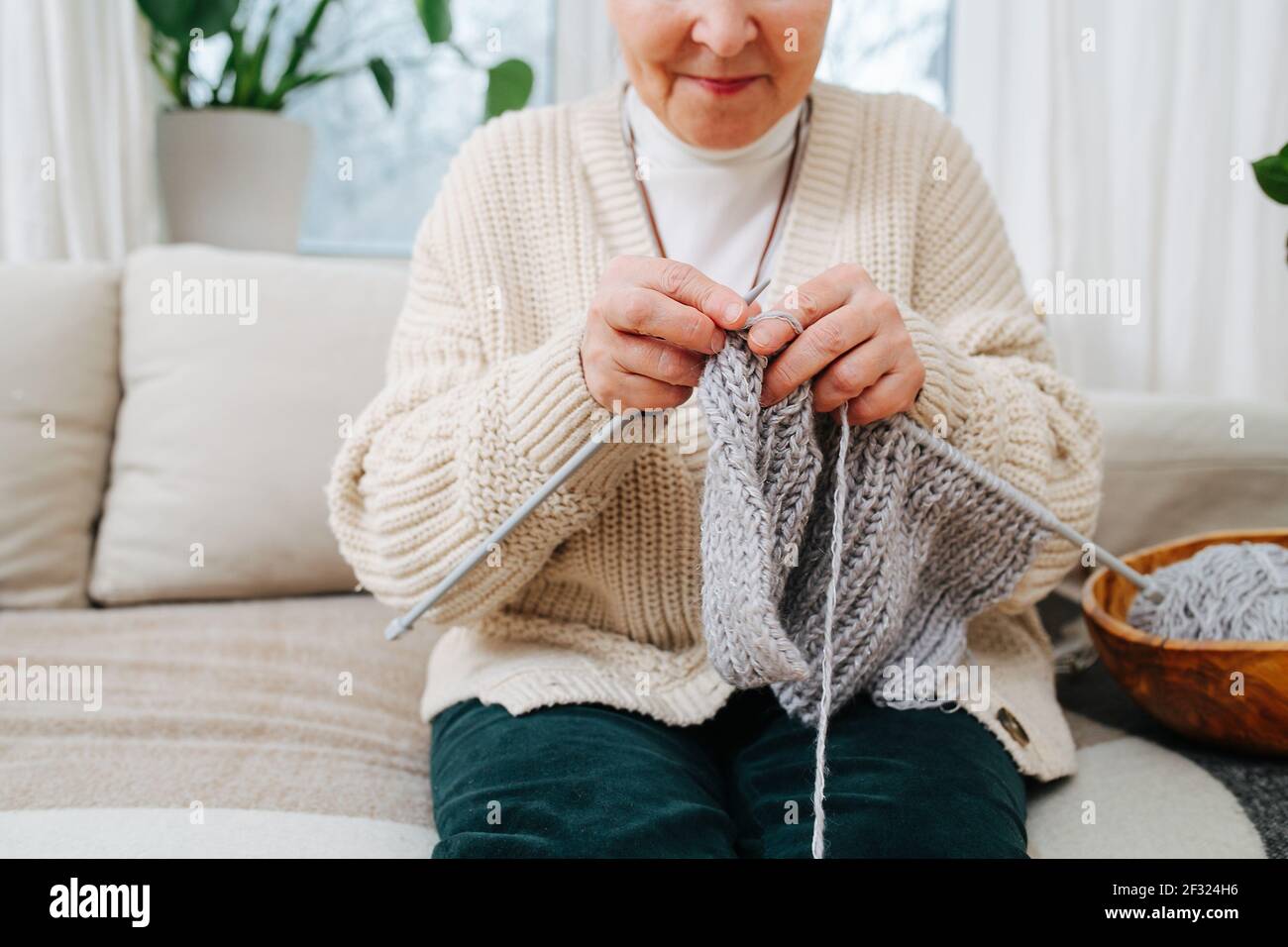 Glückliche Oma sitzt auf einer Couch zu Hause, Stricken mit Nadeln, mit grauer Wolle. Abgeschnitten, halb Gesicht. Stockfoto