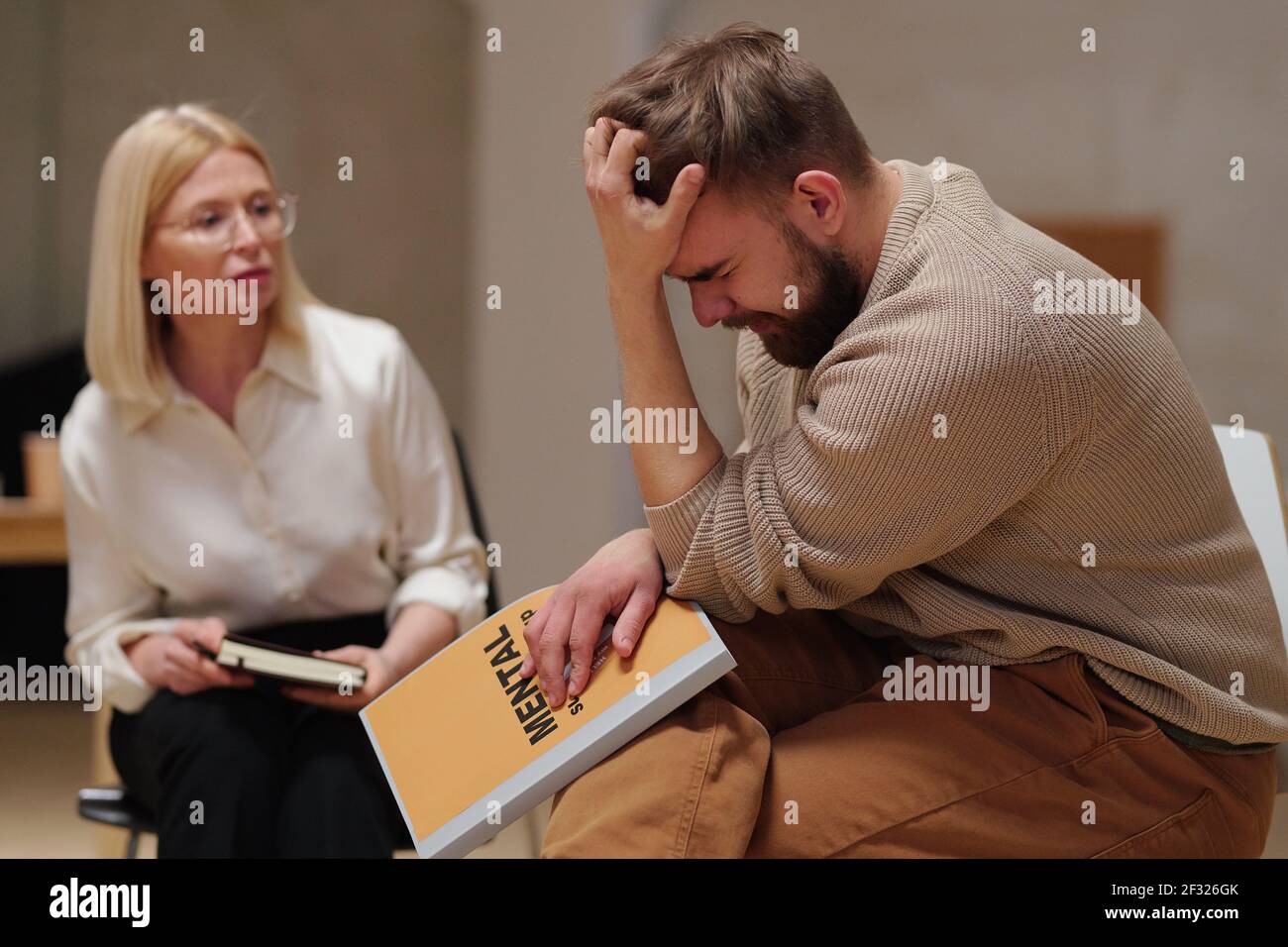 Junger bärtiger Mann mit psychologischem Problem oder Trauma hält seine Kopf in Hand, während sie von reifer blonder Beraterin sitzt Stockfoto