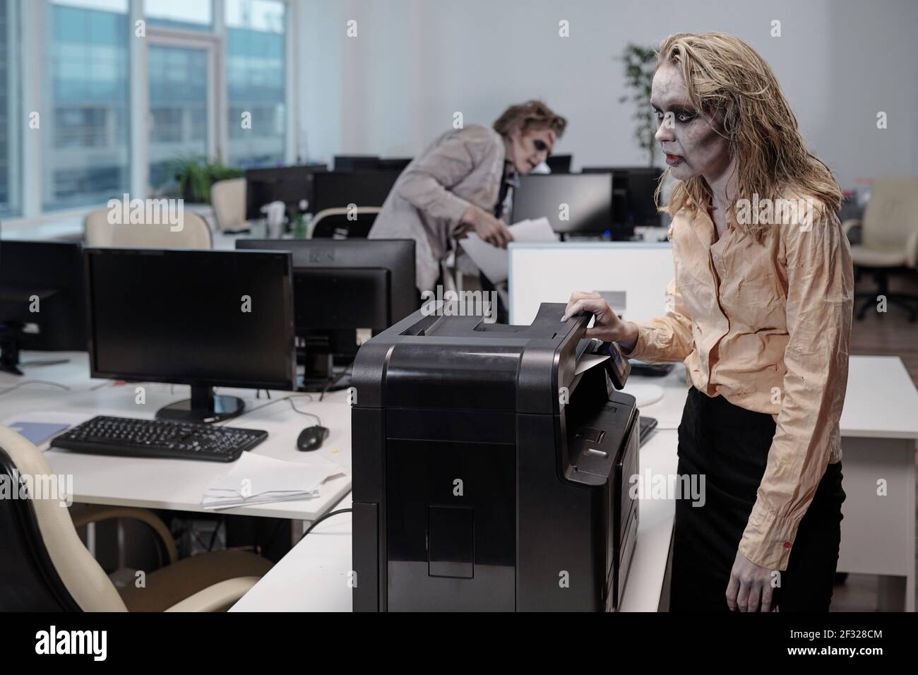 Junge Zombie Geschäftsfrau steht von xerox Maschine beim Erstellen von Kopien Von Dokumenten gegen tote männliche Kollegen mit Papieren Stockfoto