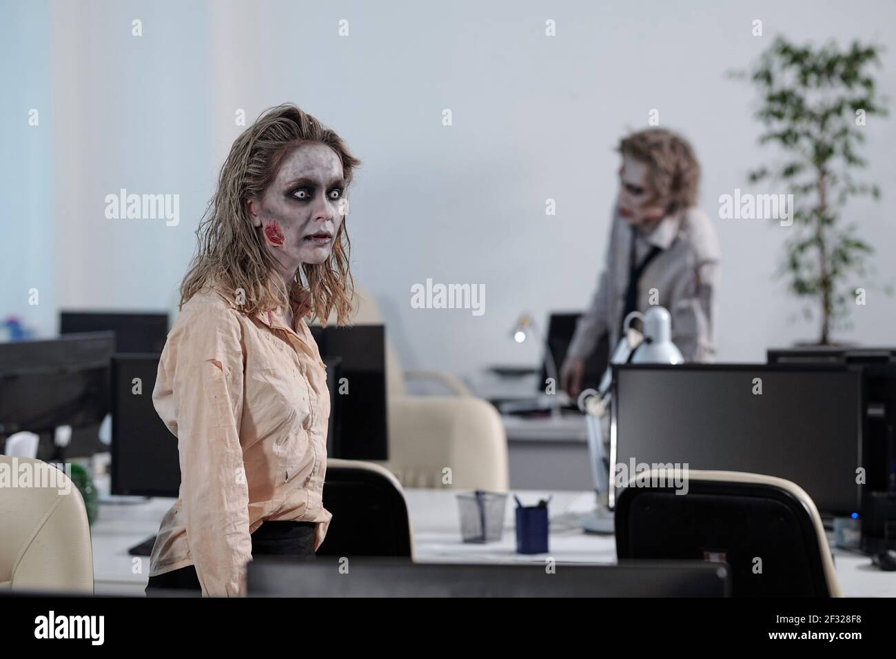 Junge Zombie Geschäftsfrau, die zwischen Schreibtischen mit Computermonitoren gegen geht Toter männlicher Kollege, der bei einem von ihnen steht Stockfoto