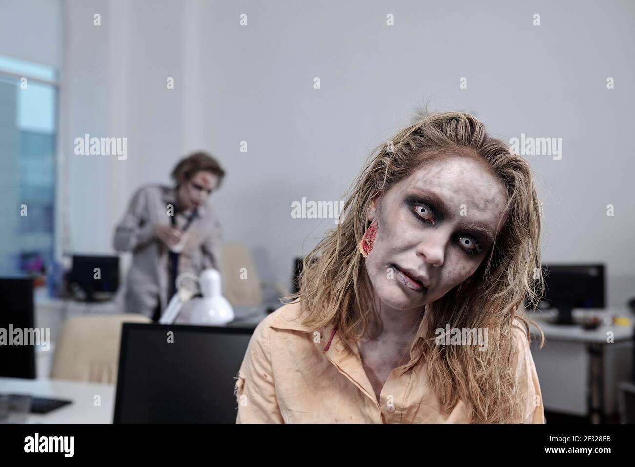 Junge Zombie Geschäftsfrau vor der Kamera auf dem Hintergrund stehen Von Schreibtischen mit Computermonitoren und toten männlichen Kollegen Stockfoto