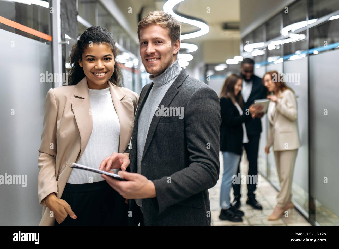 Glückliche multirassische Kollegen stehen in der Bürohalle und arbeiten zusammen an Unternehmensprojekt. Erfolgreiche Geschäftspartner blicken auf die Kamera, lächeln, Teamwork-Konzept Stockfoto