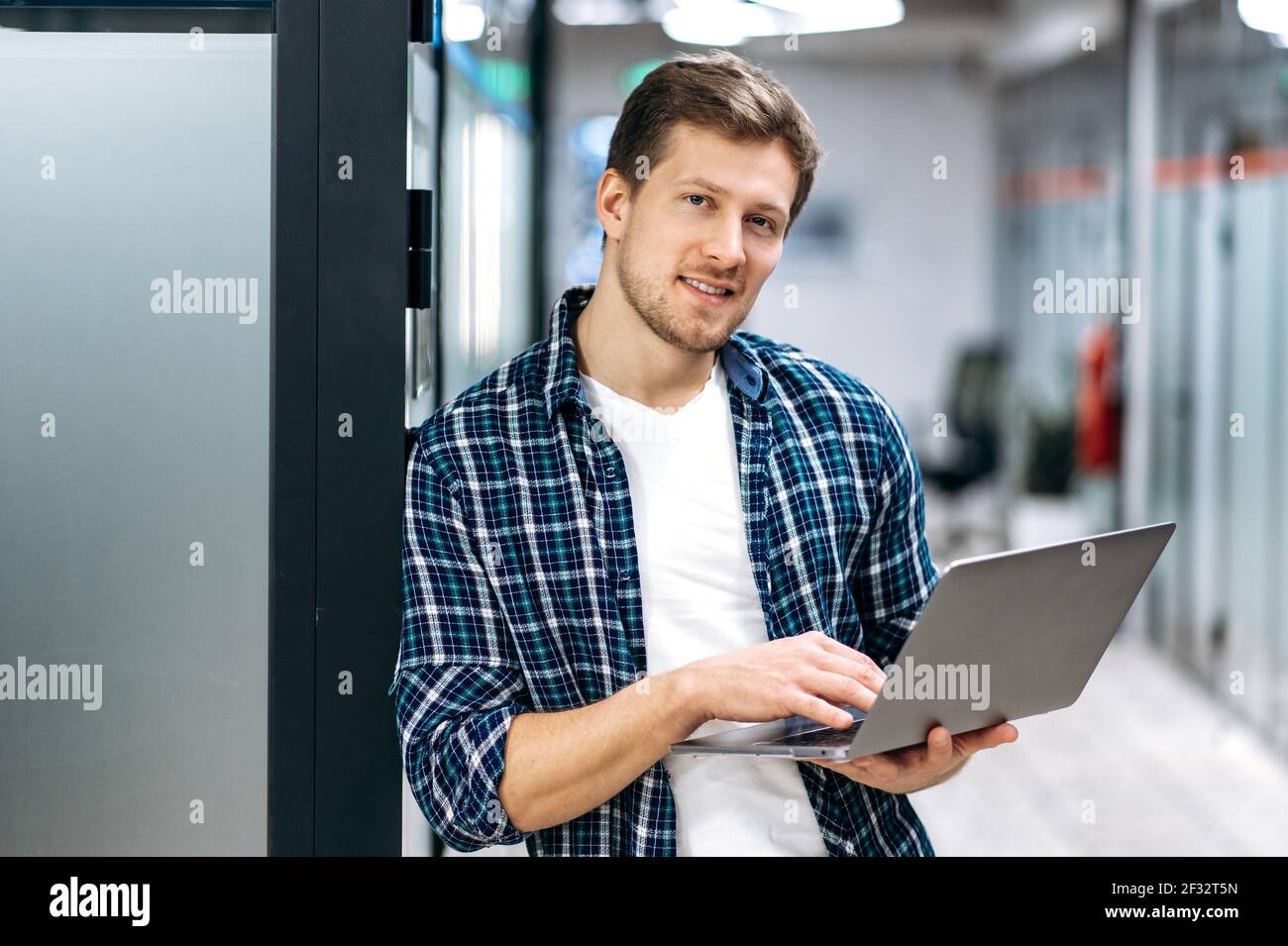 Porträt von schönen männlichen Mitarbeiter in stilvoller Kleidung am Arbeitsplatz in modernen Büro. Der junge Geschäftsmann nutzt Laptop, arbeitet an einem neuen Projekt, schaut auf die Kamera und lächelt Stockfoto