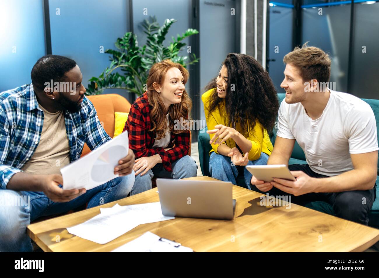 Multirassische junge Erwachsene Kollegen sitzen auf dem Sofa und diskutieren Arbeitsfragen. Fröhliche freundliche männliche und weibliche Mitarbeiter, die ein Gespräch führen, über Unternehmensprojekt sprechen, Teamwork-Konzept Stockfoto
