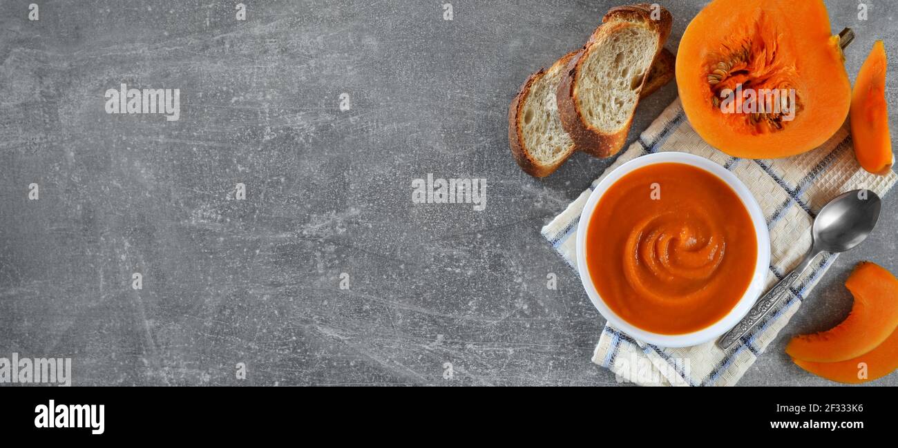 Vegetarische Kürbissuppe in einer weißen Schüssel neben frischem Kürbis und Brotscheiben auf grauem Hintergrund mit Kopierfläche, flach liegend. Gesunde Ernährung conce Stockfoto