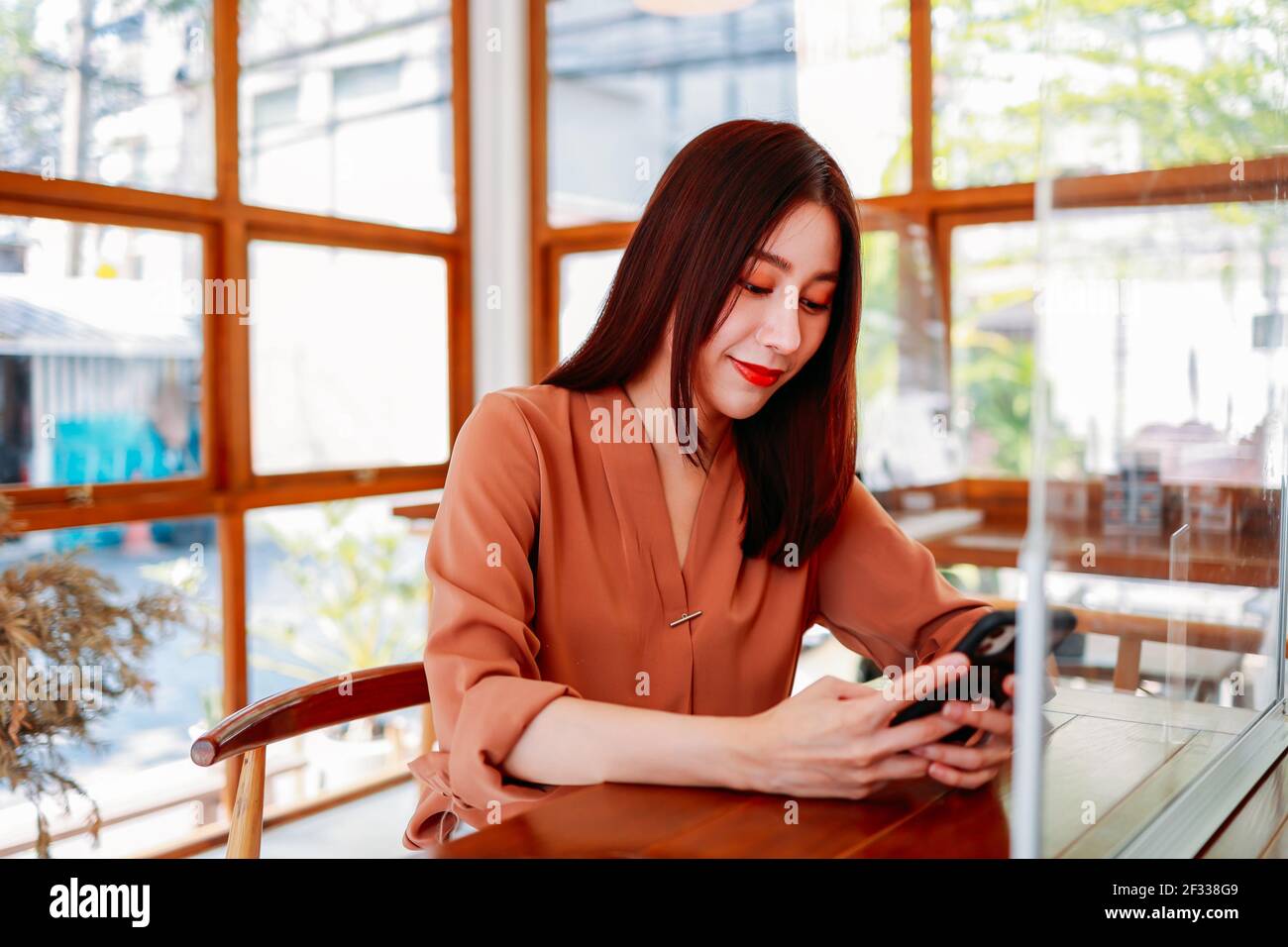 Schöne junge asiatische Frau sitzt in modernen Café für ein Entspannen Sie sich, und surfen Sie auf dem Smartphone mit Spiegel zwischen zwei Tabelle mit sozialer Distanzierung durch covid-19 Stockfoto