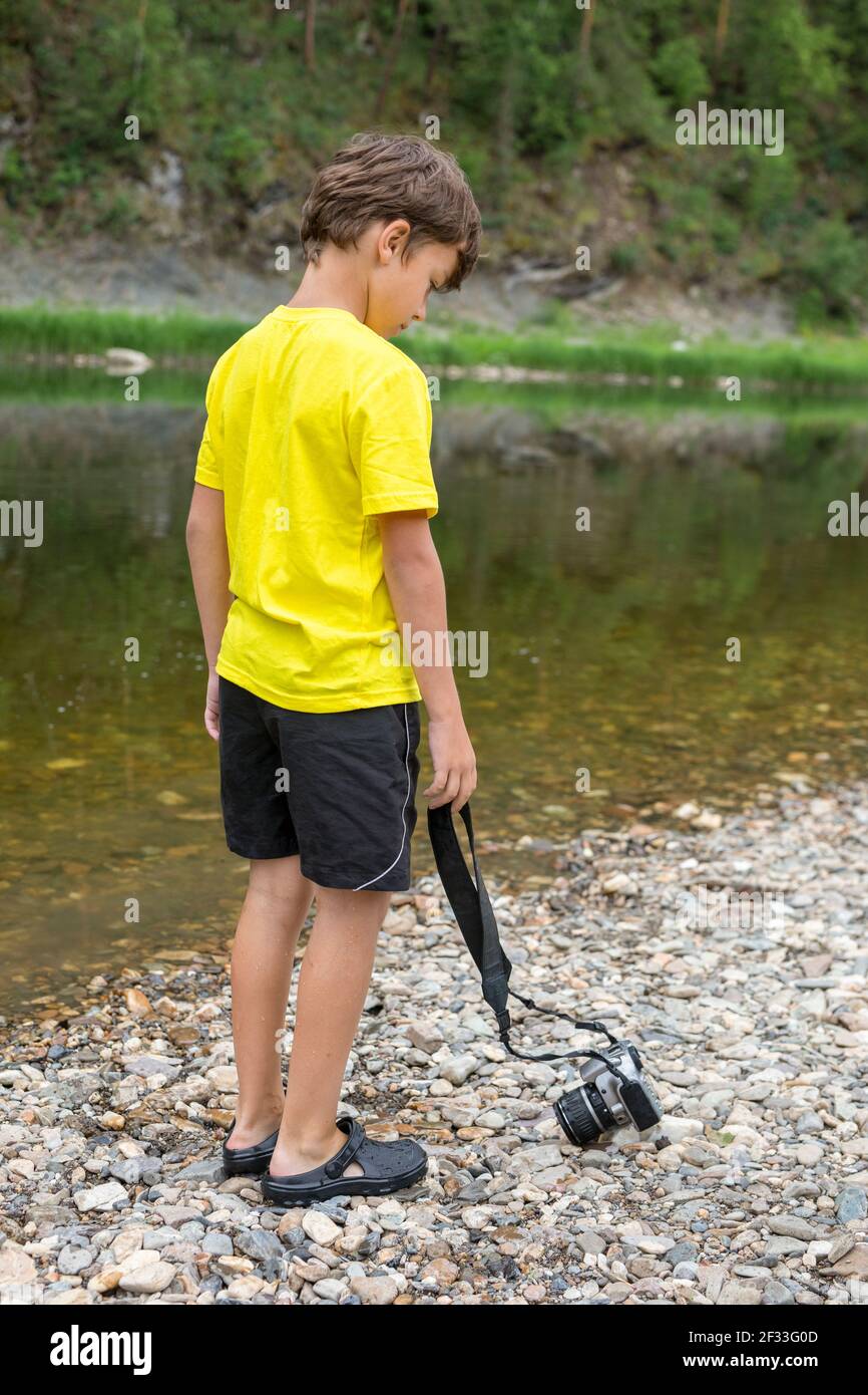 Foto eines Jungen, der zurück nach vorne steht und die Kamera auf Bachsteinen ablässt. Sommeraktivitäten für Kinder. Nachdenklichkeit. Stockfoto