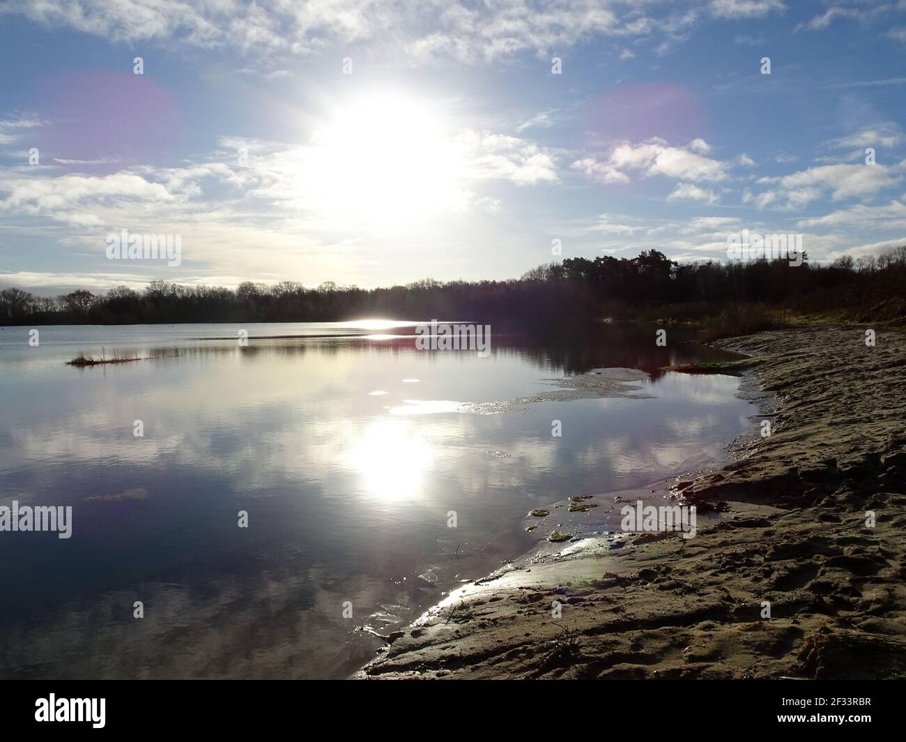 Das Ufer eines kleinen Sees. Es ist Sand. Spiegelung der Sonne im Wasser. Es ist ein heller Wintertag. Einige Streulicht. Der See hat hellblaues Wasser. Stockfoto