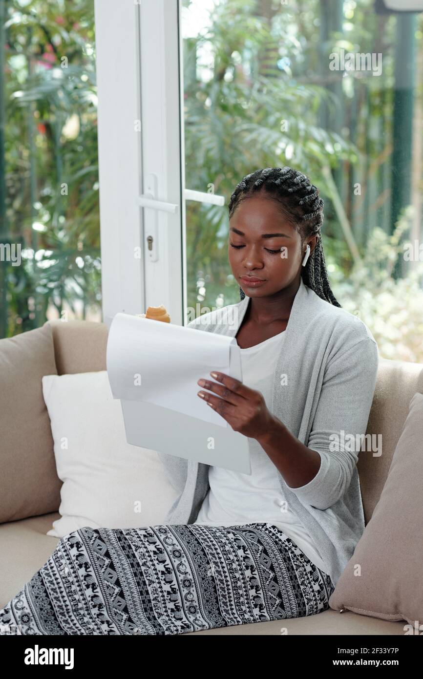 Junge schwarze Frau in Loungewear sitzen auf dem Sofa zu Hause., essen Croissant und Dokument lesen Stockfoto