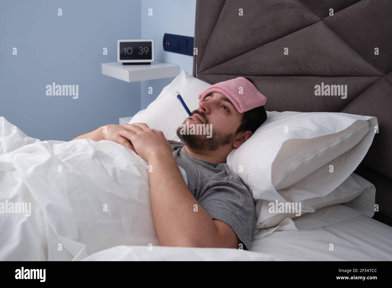 Junger latein krank im Bett mit einem Thermometer im Mund und einem Tuch auf dem Kopf. Digitale Uhr im Hintergrund. Stockfoto