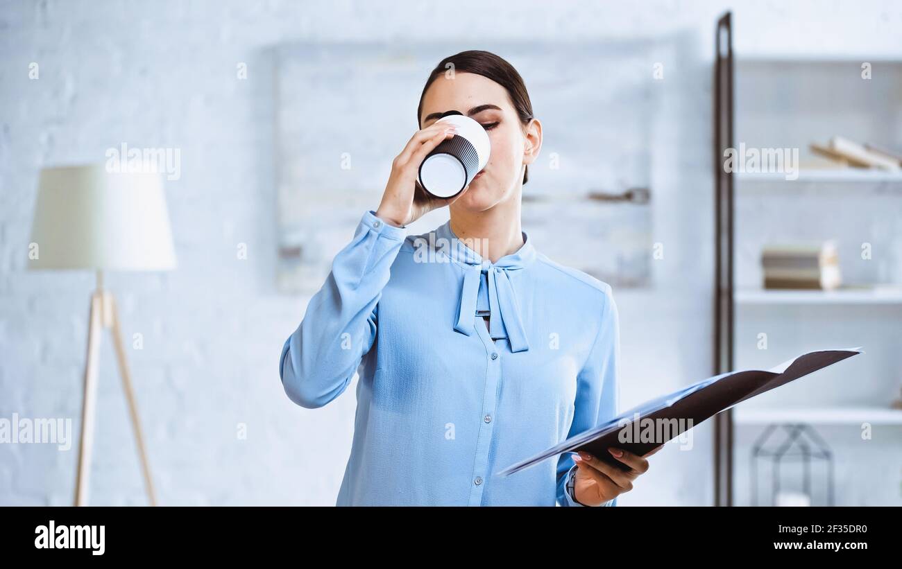 Junge Geschäftsfrau trinkt Kaffee, während sie Ordner mit Dokumenten in Büro Stockfoto