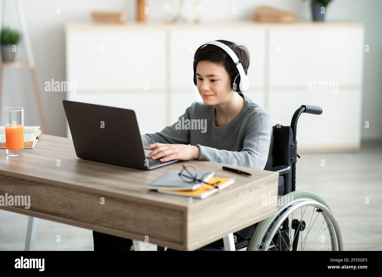 Behinderter Teenager Junge, der Musik hört, während er Online-Hausaufgaben auf dem Laptop macht, drinnen Stockfoto