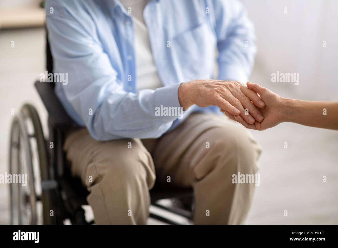 Ältere behinderte Menschen unterstützen. Junge Frau, die die Hand eines behinderten älteren Mannes im Rollstuhl in Innenräumen hält, Nahaufnahme Stockfoto
