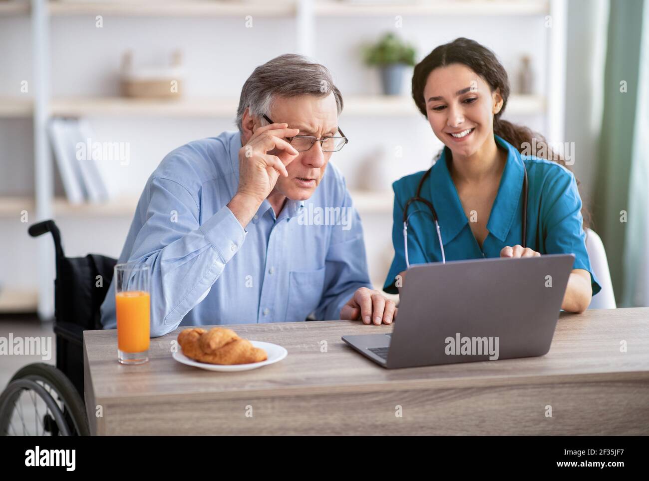 Junge Krankenschwester zeigt älteren behinderten Mann, wie man Laptop zu verwenden Computer im Altersheim Stockfoto