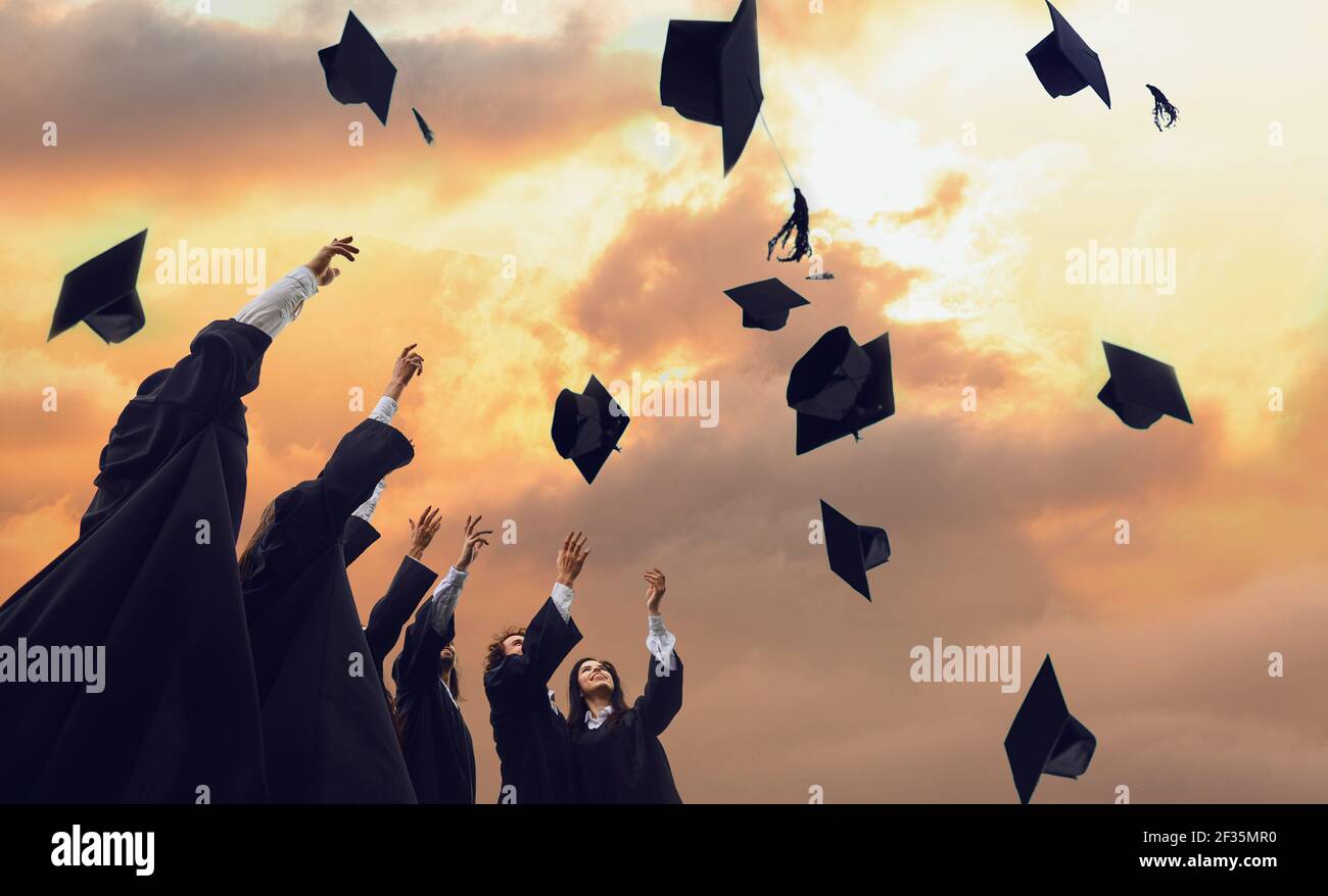 Absolventen in schwarzen Gewändern werfen ihre akademischen Hüte vor dem Hintergrund eines magischen Sonnenuntergangs. Stockfoto