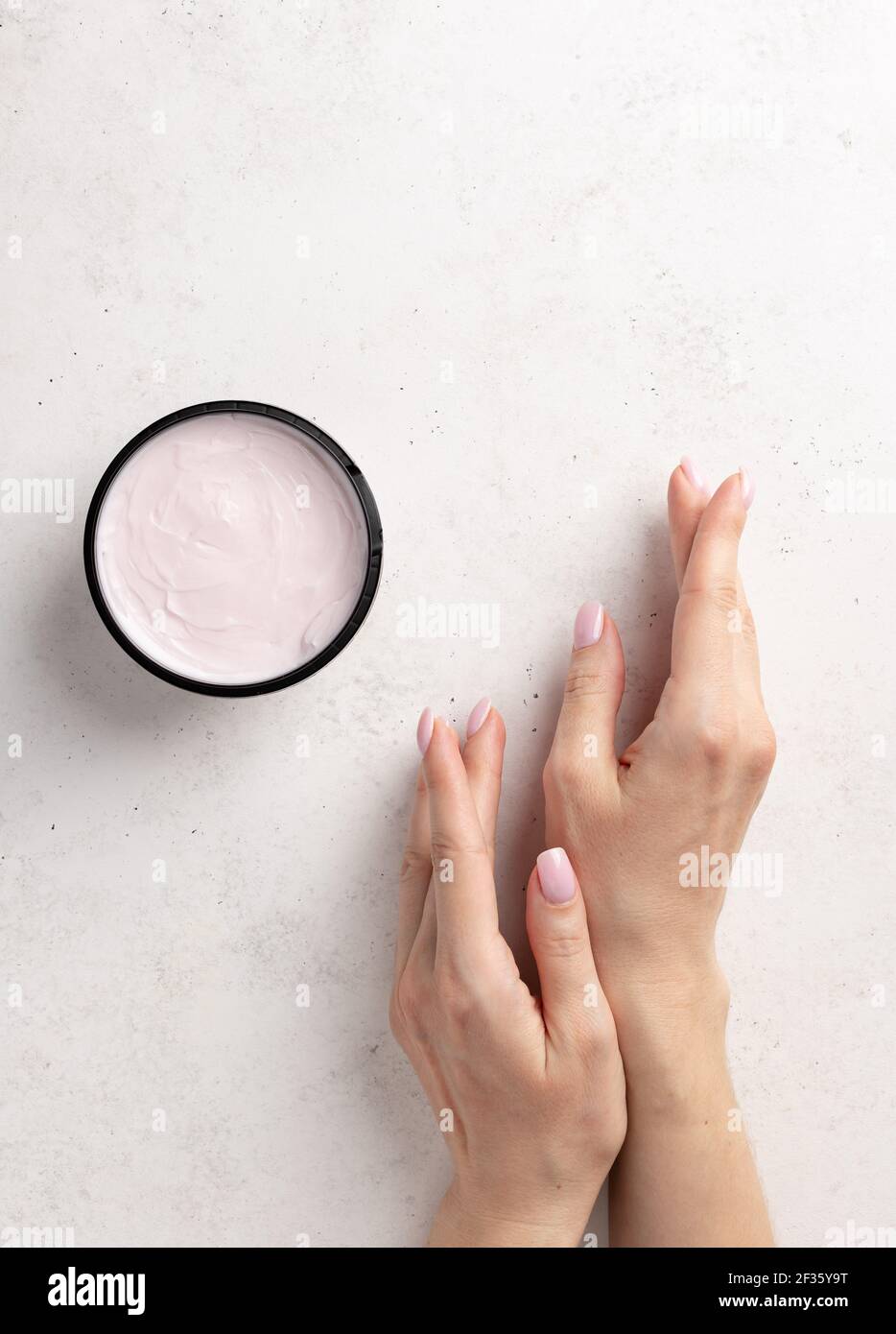 Gepflegte Frauenhände mit einer zarten rosa Maniküre und Creme-Glas auf einem weißen Stein Hintergrund. Minimalismus. Hautpflege. Vertikales Bild. Draufsicht. Ort Stockfoto