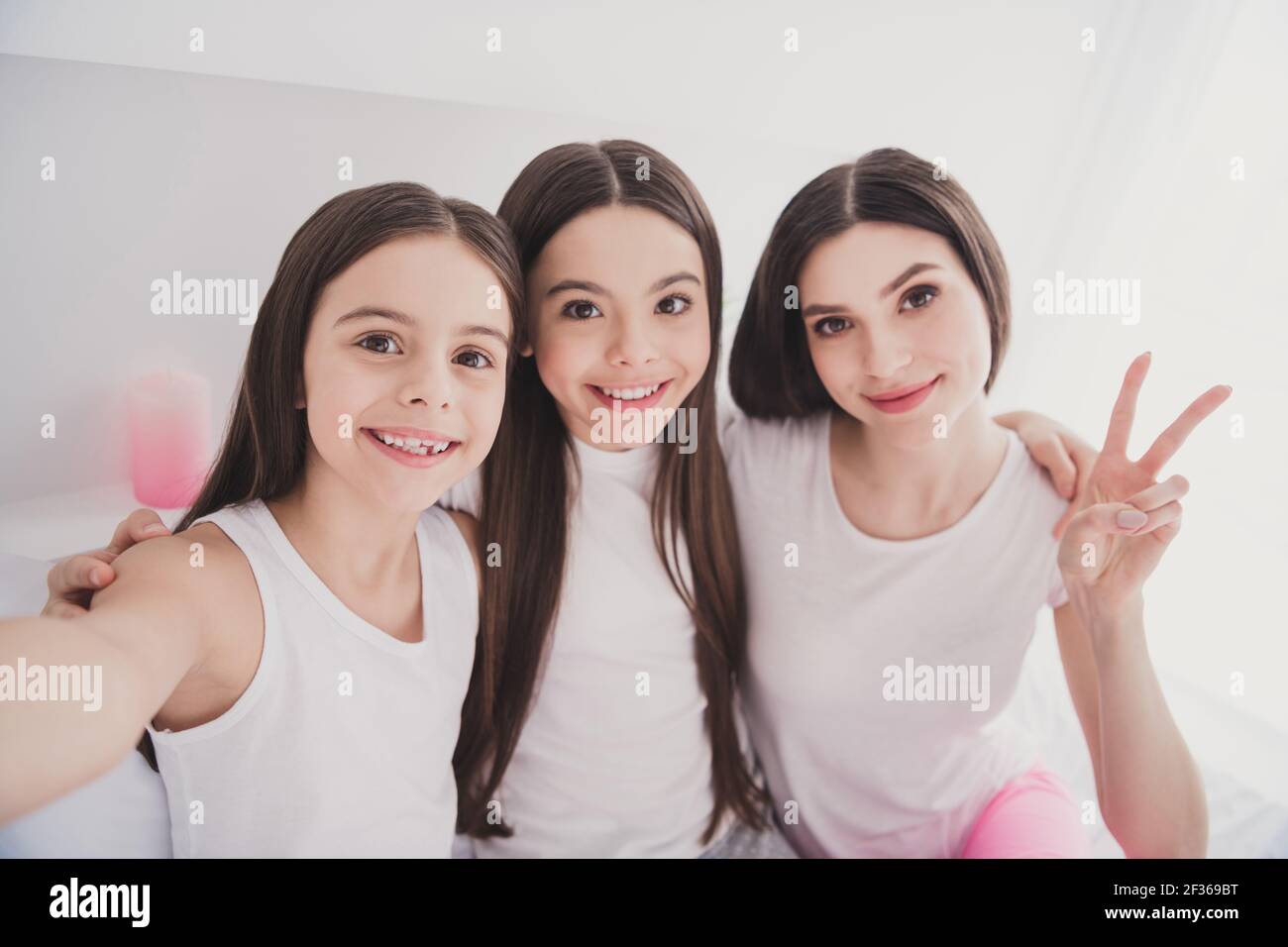 Foto von süßen lustigen drei Geschwister Nachtwäsche sitzend Bett lächelnd Umarmend tacking Selfie zeigt V-Zeichen drinnen im Zimmer zu Hause Stockfoto