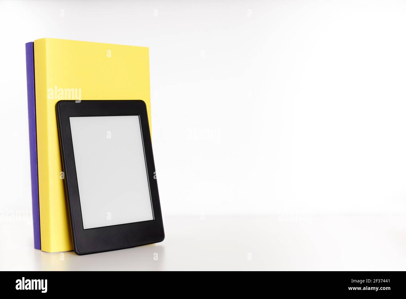 Gelbe und lila Bücher mit leerem Rücken und einem elektronischen Leser auf einem weißen Regal mit weißem Hintergrund. Konzepte der Technik, des Lesens und der Pädagogik Stockfoto