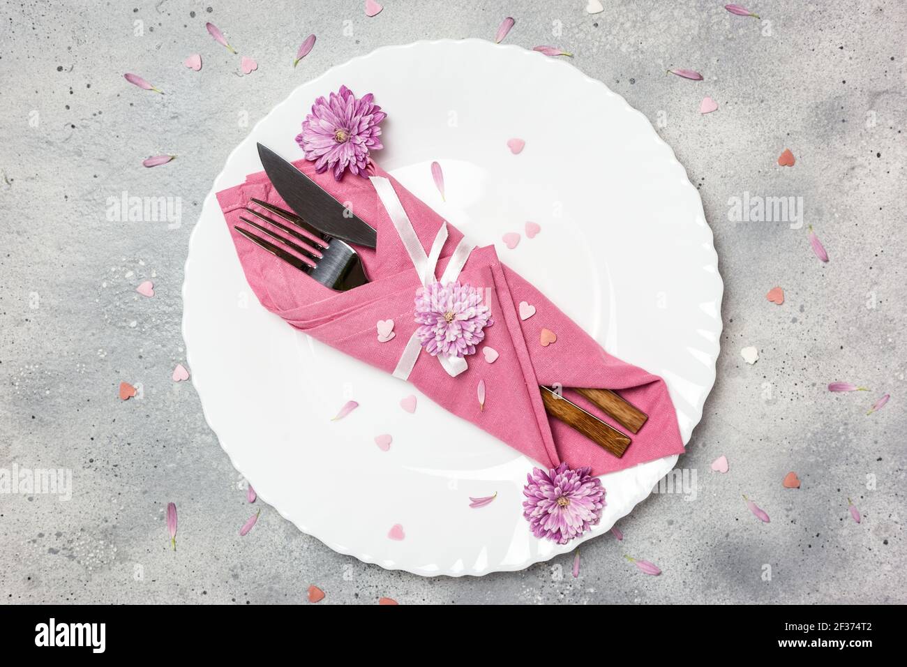 Schöne festliche Tischdekoration mit weißem Teller, Serviette, Blumen und Herzen auf grauem Steinhintergrund. Draufsicht mit Kopierbereich. Stockfoto